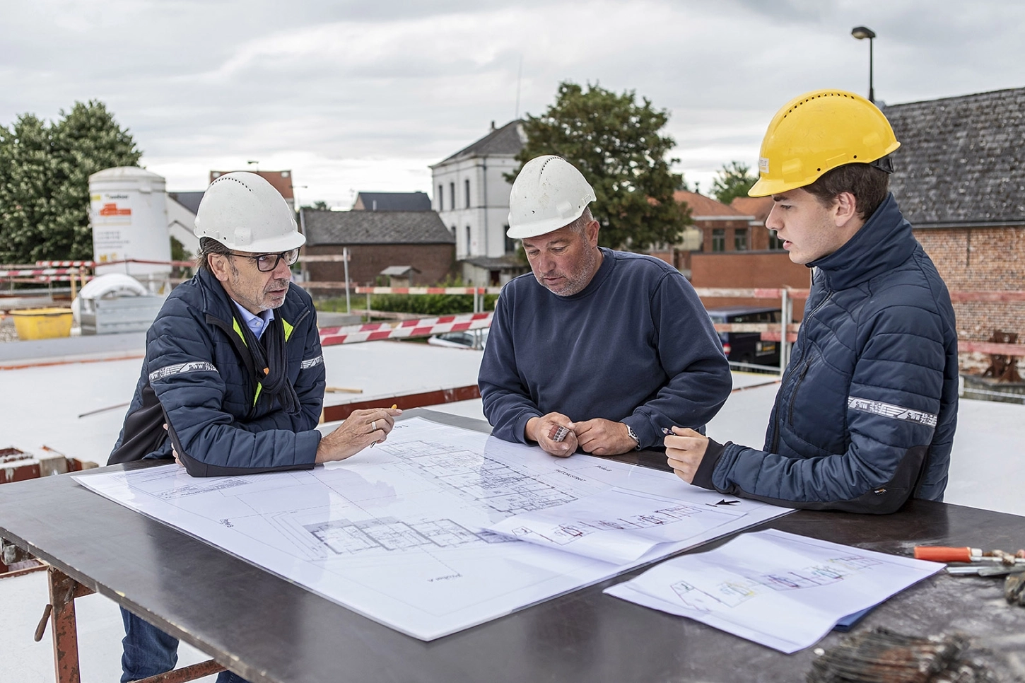 Drie ervaren medewerkers staan rond een tafel op een werf en bekijken de bouwplannen.